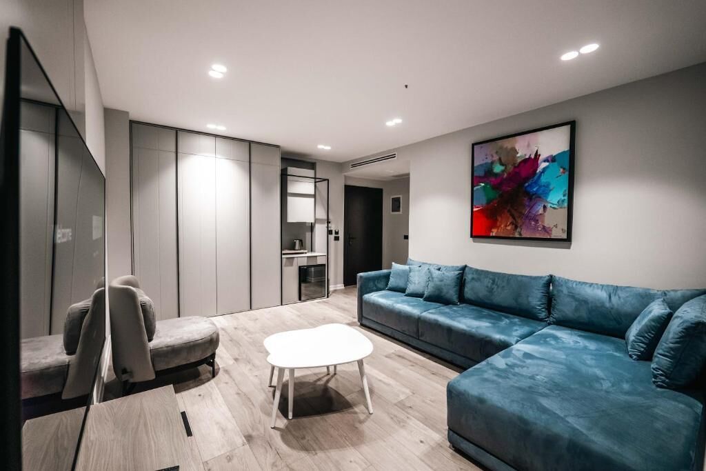 A modern living room with a blue sofa, a white coffee table, and a grey armchair. There is a kitchen area in the background with cabinets and a countertop.