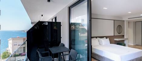 A modern bedroom with a large bed, a glass door leading to a balcony with a table and chairs, and a view of the sea.