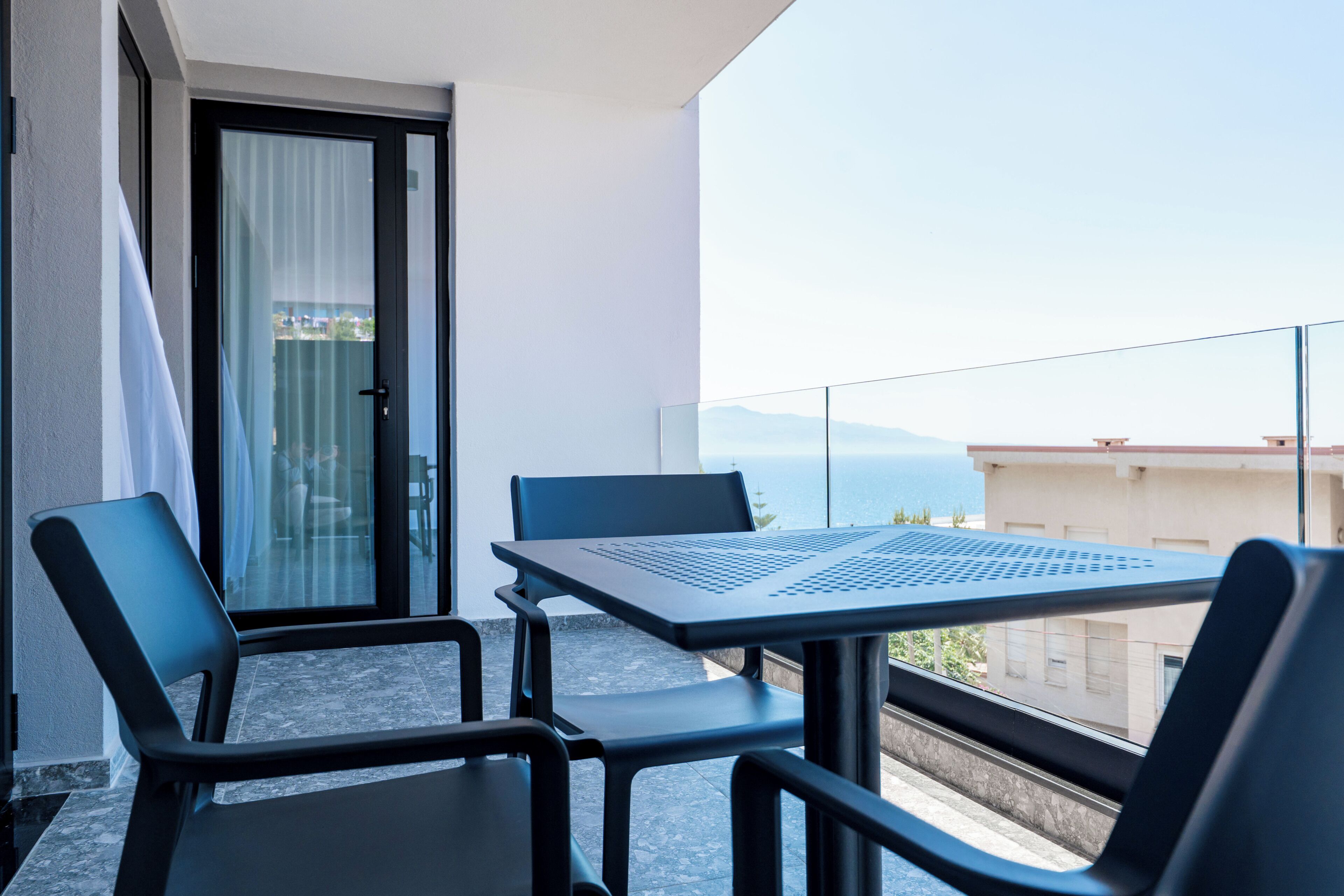 A balcony with a table and chairs, a sliding glass door, and a view of the sea and buildings.