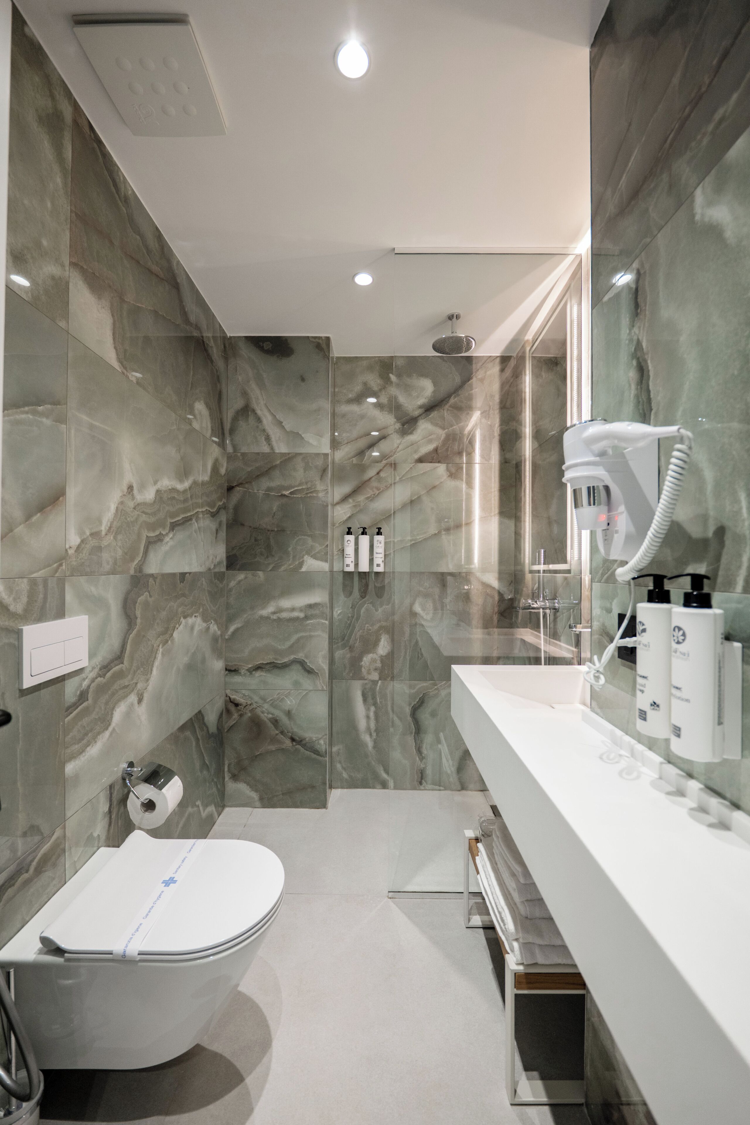 Ein modernes Badezimmer mit Marmorwand, Toilette in Weiß, Duschabtrennung aus Glas und Waschtisch mit Pflegeprodukten.