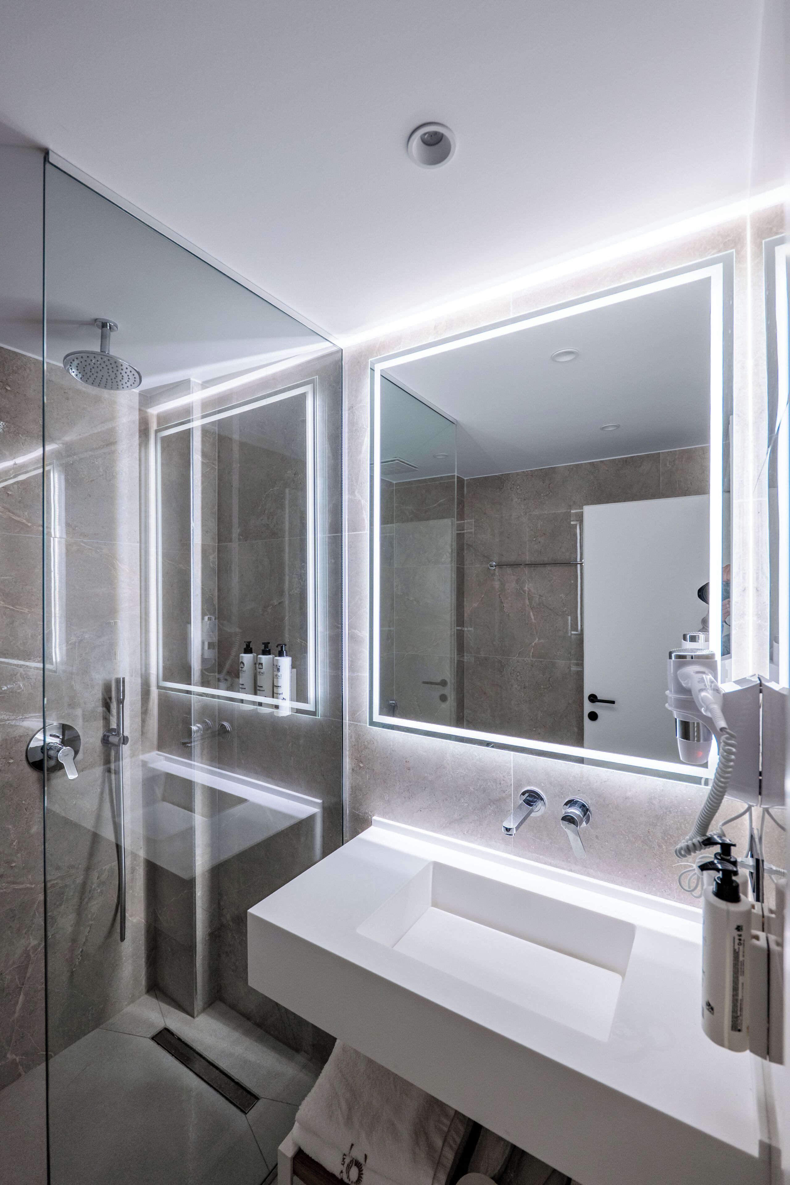 Een moderne badkamer met een douchecabine van glas, een witte wastafel en twee spiegels met ingebouwde verlichting.
