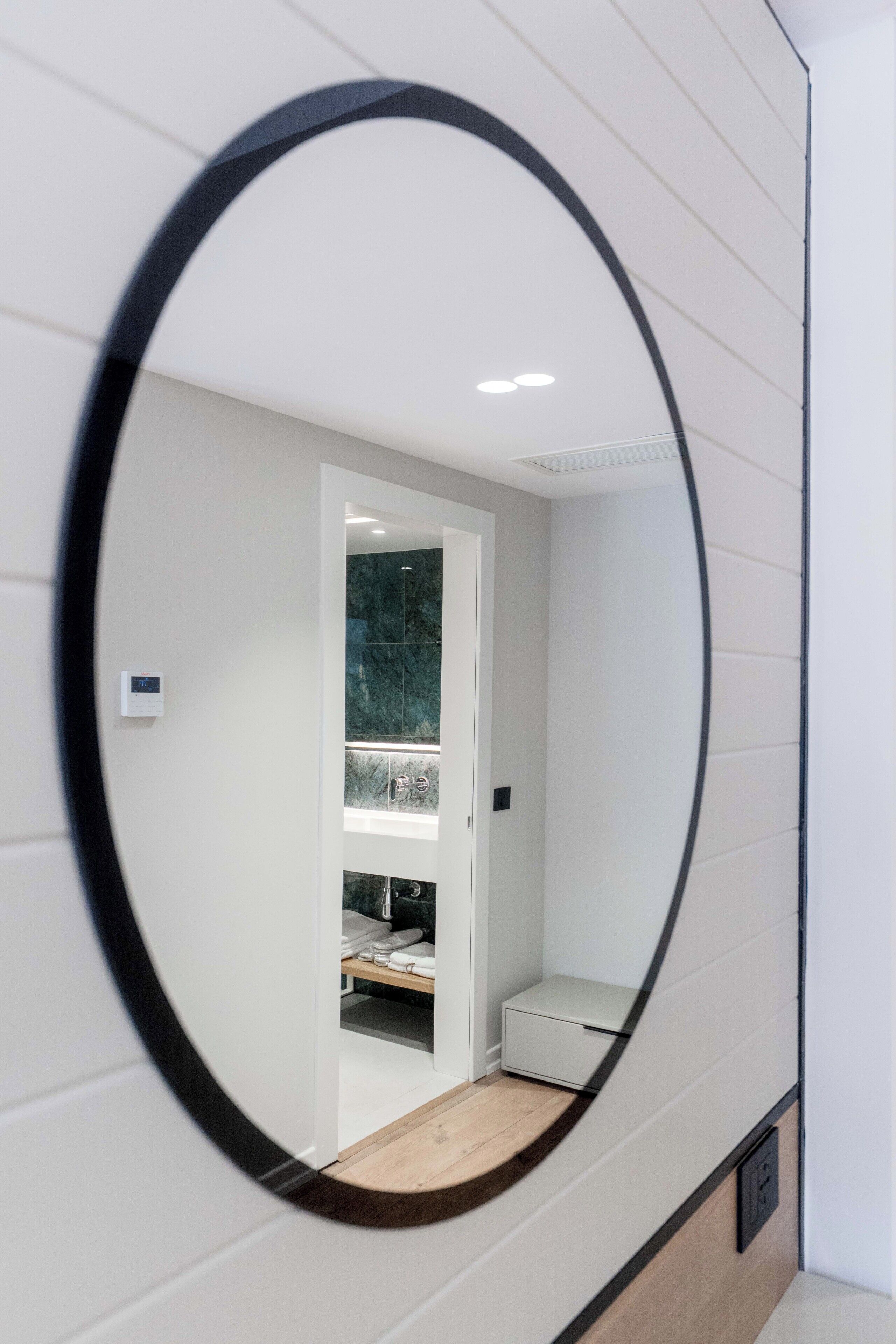 A mirror reflecting a room with a white door, a shelf with items, and a small white cabinet.