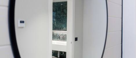 A mirror reflecting a room with a white door, a shelf with items, and a small white cabinet.