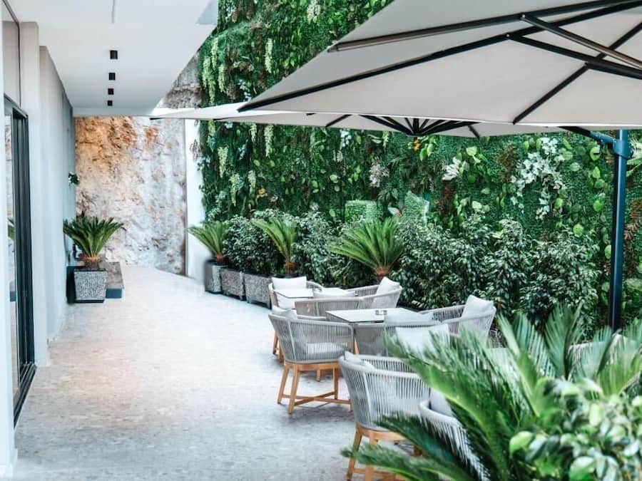 Un patio interior con pared de piedra, plantas verdes y mobiliario exterior bajo un gran paraguas.