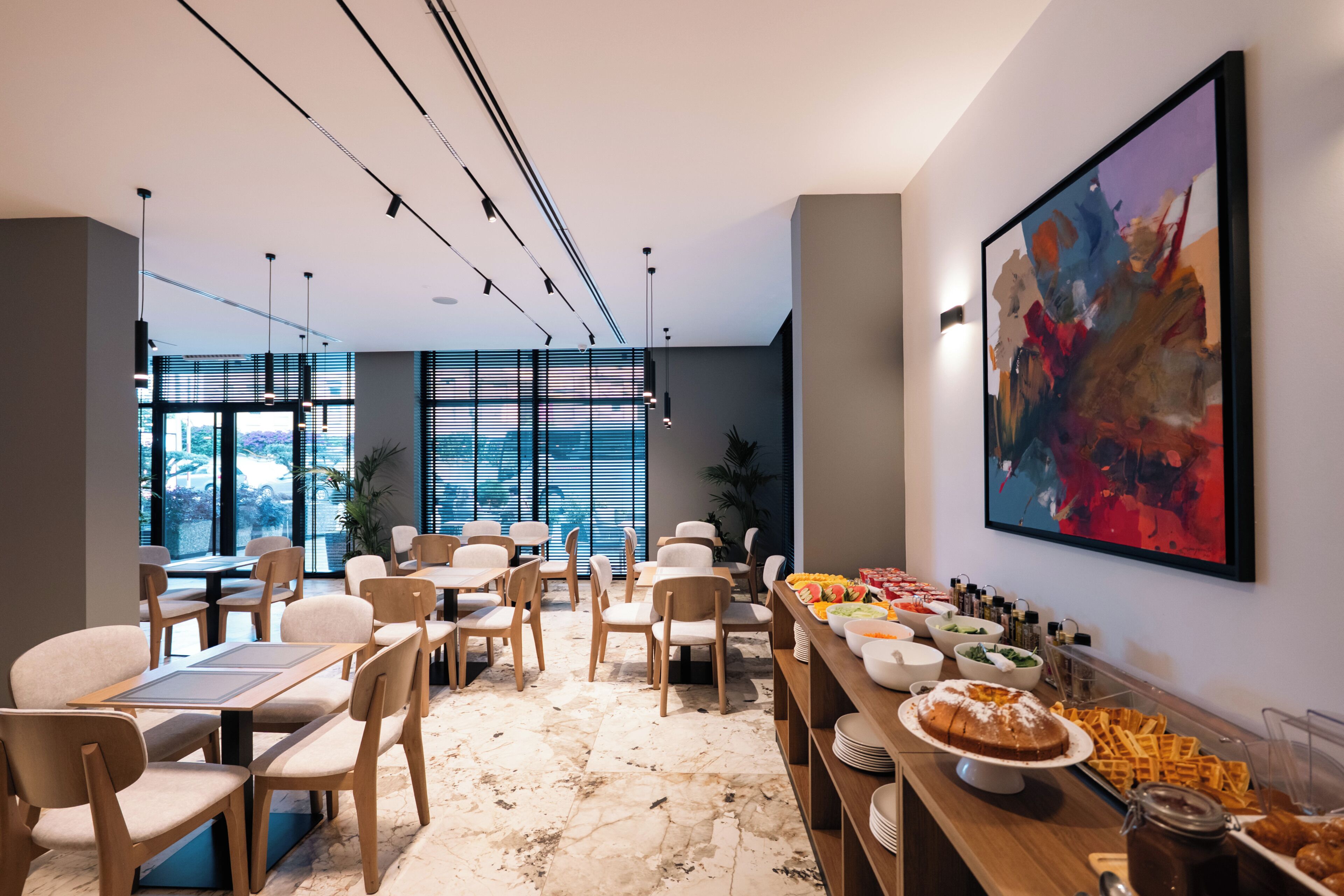 A modern dining area with tables and chairs, a buffet with various food items, and a large abstract painting on the wall.