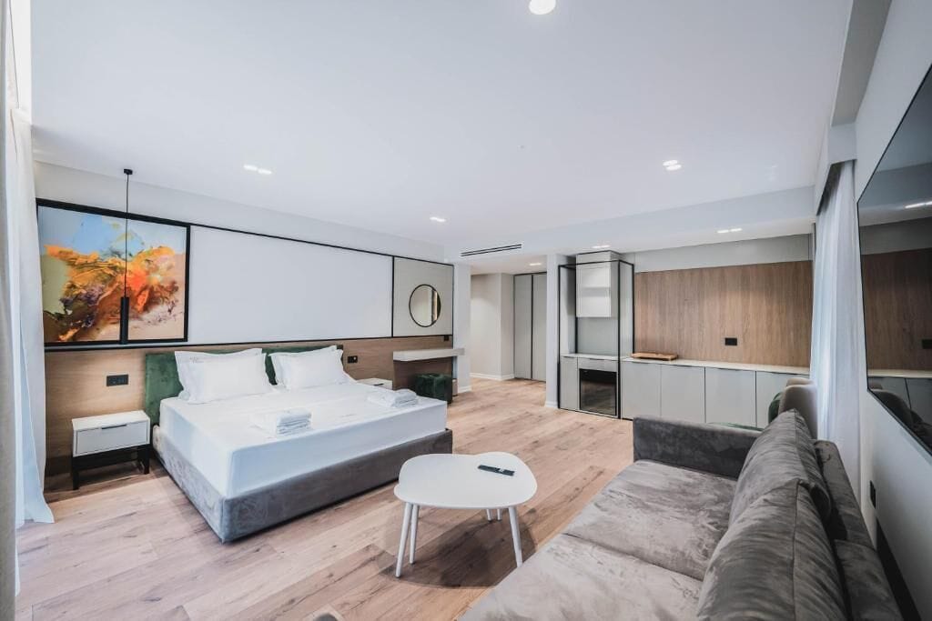 Una habitación de hotel moderna con una cama grande, una mesita pequeña, un sofá y una zona de cocina con armarios.