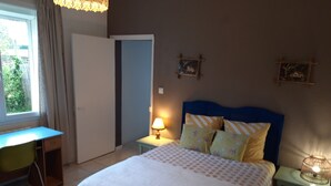 Ein Schlafzimmer mit Bett, Schreibtisch und Stuhl. Eine Tür, ein Nachttisch mit Lampe und eine Wand mit zwei gerahmten Bildern sind ebenfalls vorhanden.