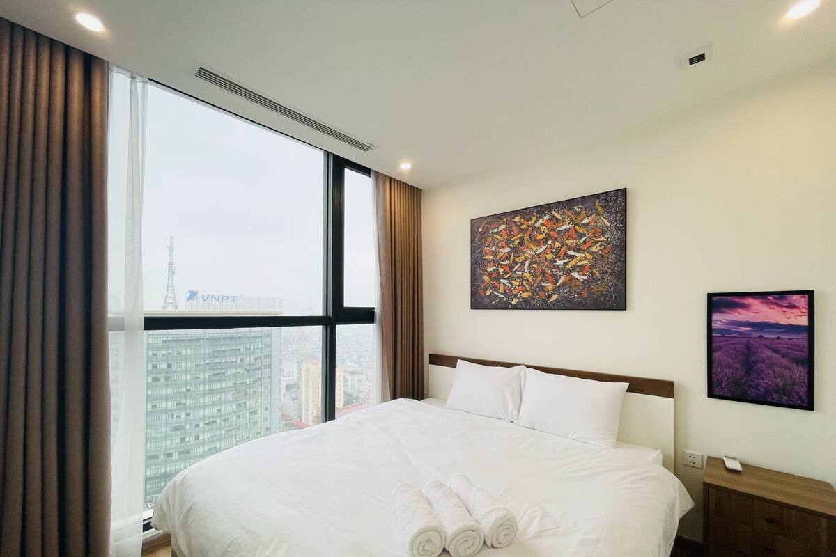 A hotel room with a large bed, a nightstand, a wall-mounted painting, and a window with curtains.