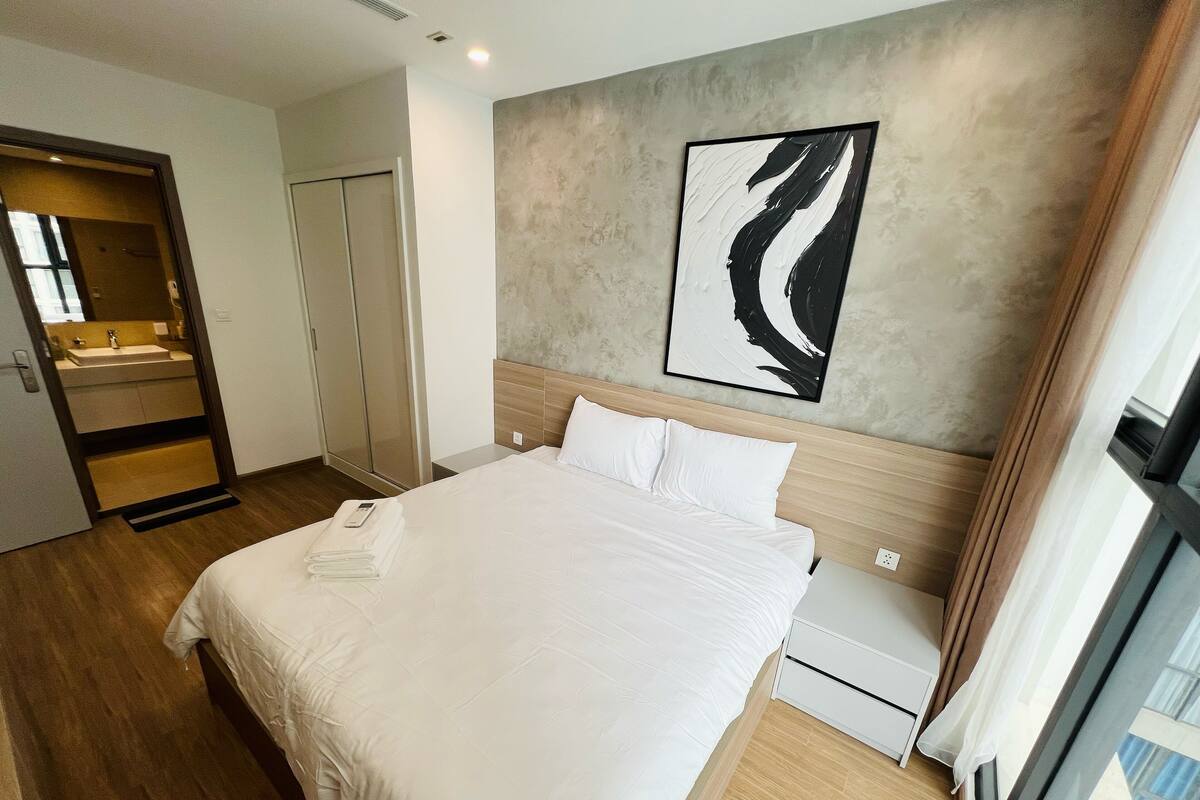 A hotel room with a large bed, a wooden headboard, a modern abstract painting on the wall, and a view of the city through the window.