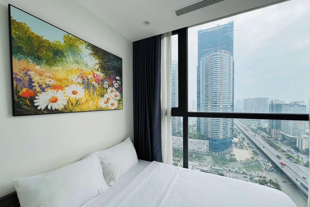 A hotel room with a large bed, a cityscape view through the window, and a floral painting on the wall.