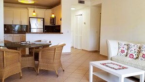 A dining area with wicker chairs and a glass table, a kitchenette with stainless steel appliances, and a living area with a sofa and patterned cushions.