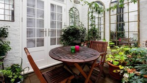 A round wooden table with chairs, surrounded by potted plants and a white brick wall. - The Westminster Courtyard Apartment, by Veeve (London)