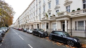 A row of white townhouses with numbered doors, parked cars, and autumn leaves on the street. - The Westminster Courtyard Apartment, by Veeve (London)