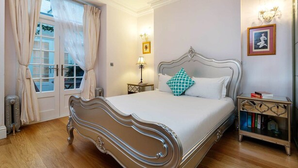 A bedroom with a large bed, a chandelier, a bedside table with books, and a window with curtains. - The Westminster Courtyard Apartment, by Veeve (London)