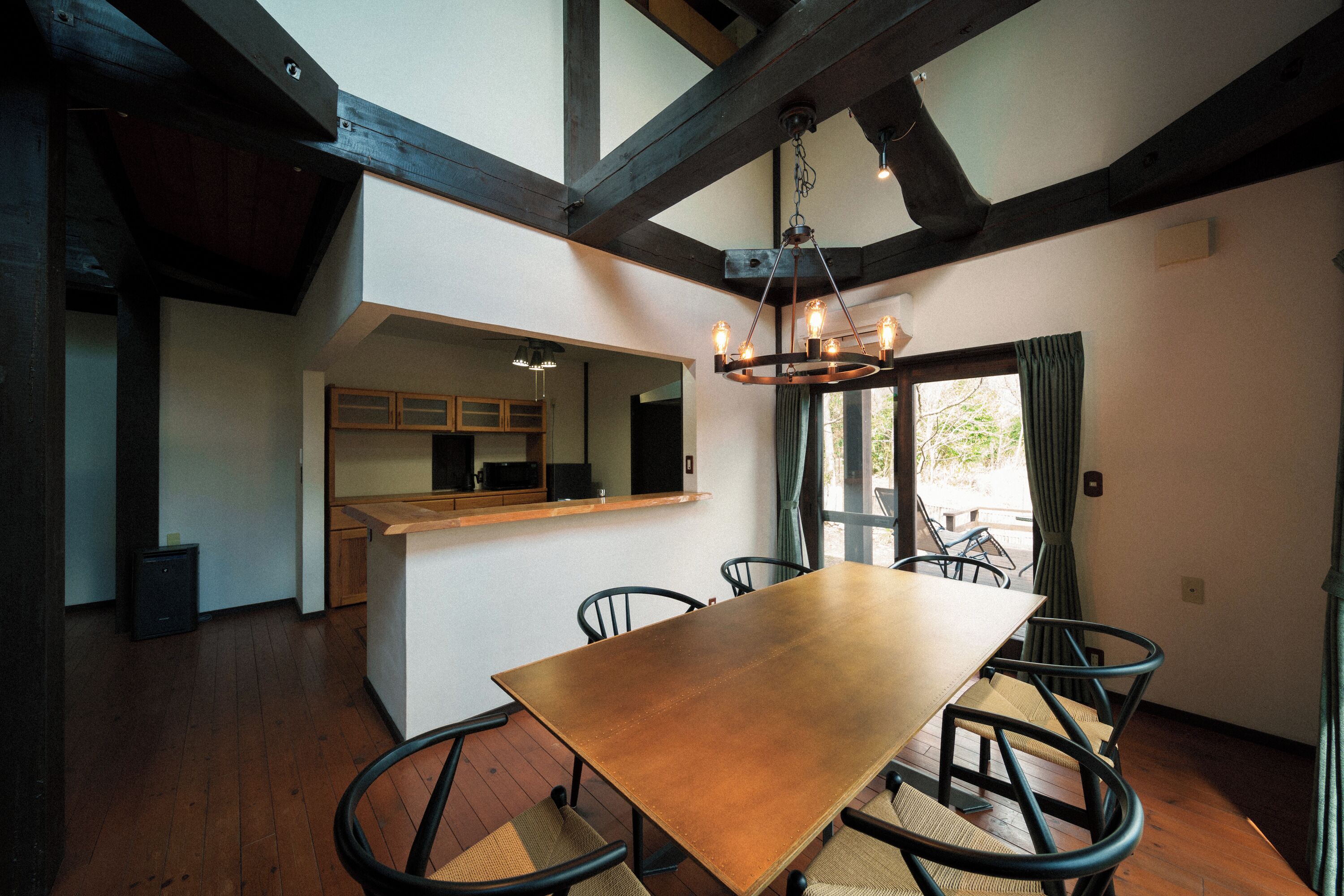 A dining area with a wooden table and chairs, a kitchen in the background, and a large window with curtains.