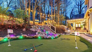 A well-lit mini-golf course with numbered holes, a wooden staircase, and a house in the background. - Asheville Mansion: Spa, Games, Sleeps 28 (Fletcher)