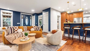 A modern living room with a sofa, coffee table, and a small round table. There is a kitchen area with wooden cabinets and a bar stool. - Asheville Mansion: Spa, Games, Sleeps 28 (Fletcher)