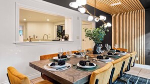 A dining area with a long wooden table set for six, surrounded by cushioned chairs. A large mirror reflects the room, and a wooden slatted ceiling adds a modern touch. - Lux Nature Villa |Hot Tub + Theater + Games + Spa! (Fletcher)