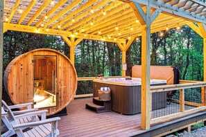 A wooden cabin with a hot tub and deck chairs, surrounded by trees.