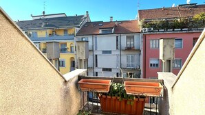 A small balcony with potted plants, a hose, and a window overlooking neighboring buildings.