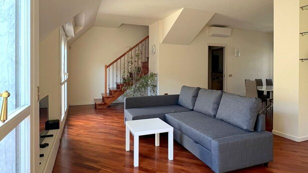 A spacious living room with a grey sectional sofa, a white coffee table, and a wooden floor. A staircase with a wooden railing leads to an upper level.