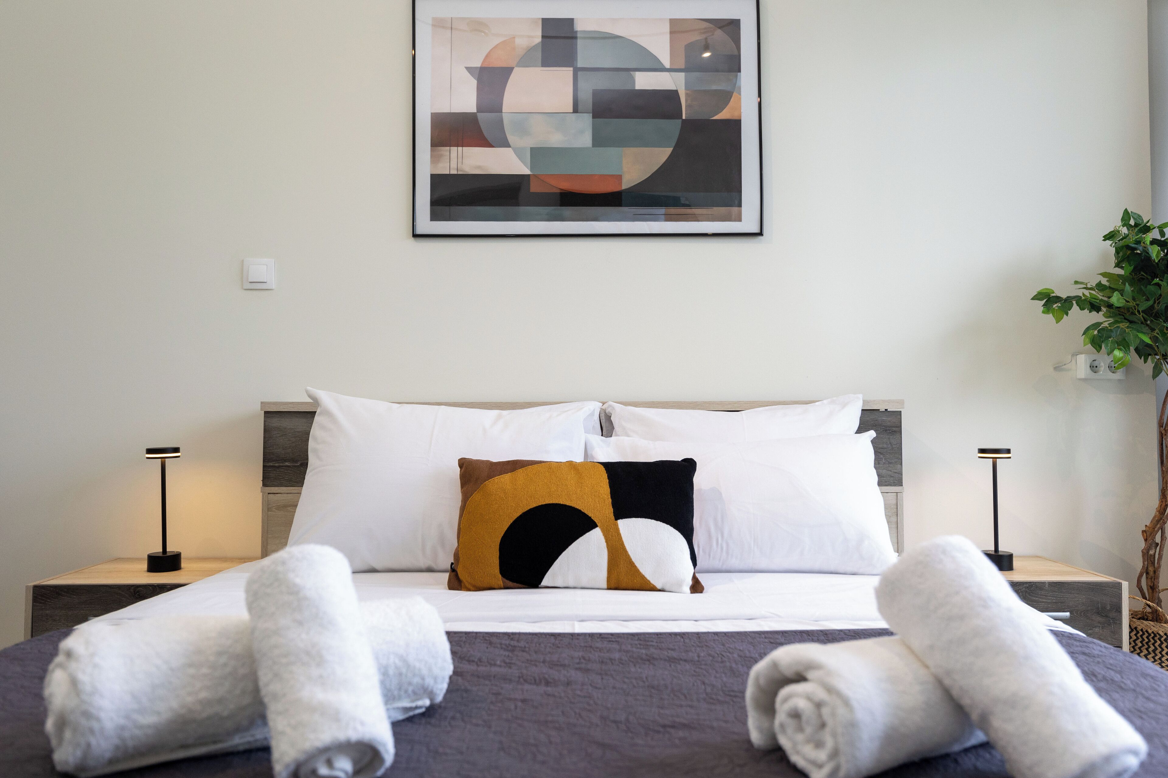 Ein ordentlich bezogenes Bett mit weißen und braunen Kissen, zwei Nachttischlampen und einem gerahmten, abstrakten Kunstwerk darüber.