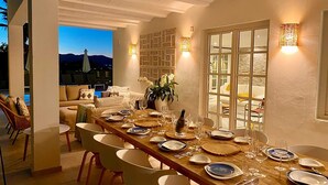 Un comedor con una larga mesa dispuesta para una comida, rodeada de sillas, y vistas a las montañas al fondo.