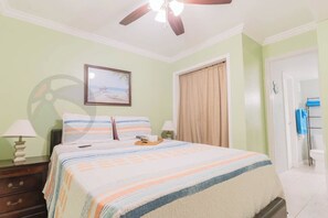 Ein Schlafzimmer mit Bett, Deckenventilator, Nachttisch, Lampe, gerahmtem Bild an der Wand und einer Badezimmertür.