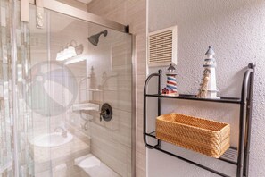 Une salle de bain avec une douche à parois de verre, un lavabo blanc et une étagère en métal noir sur laquelle repose un panier en osier.
