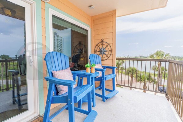 Un balcon avec des fauteuils Adirondack bleus, une petite table sur laquelle sont posés un ananas et une plante, et une décoration en forme de boussole accrochée au mur.