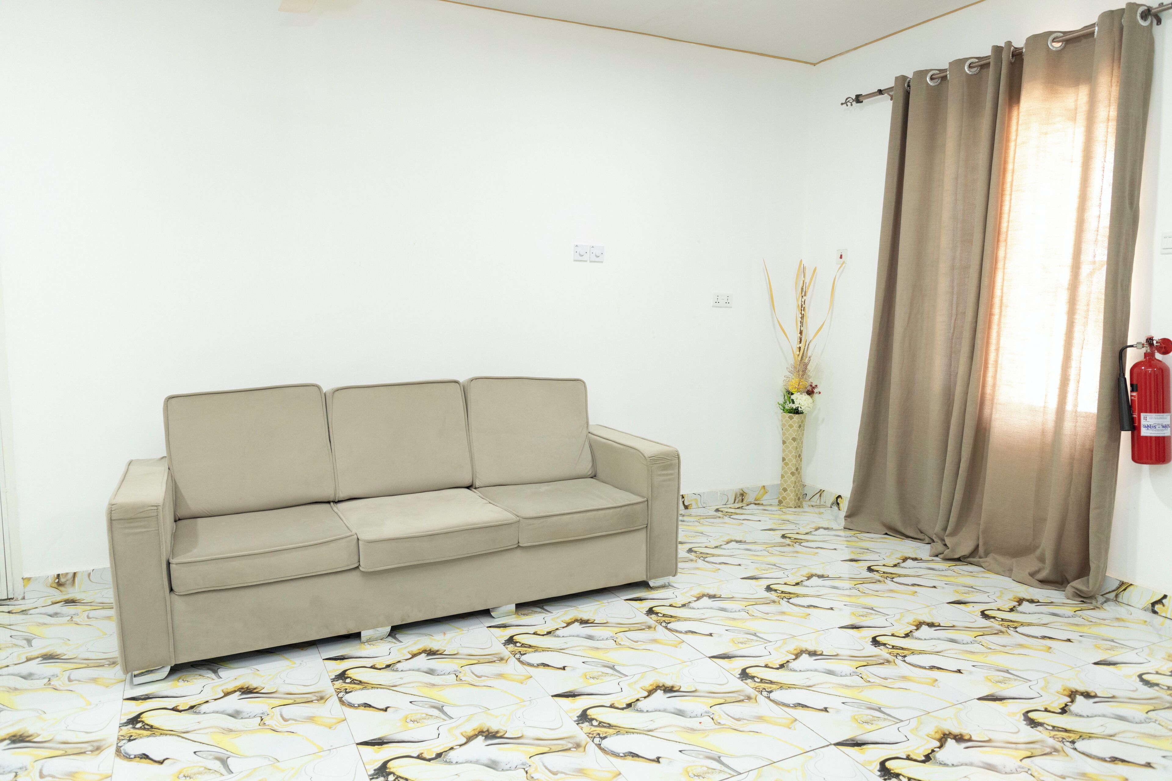 Una habitación con un sofá color beige, piso estampado, cortina y extintor.