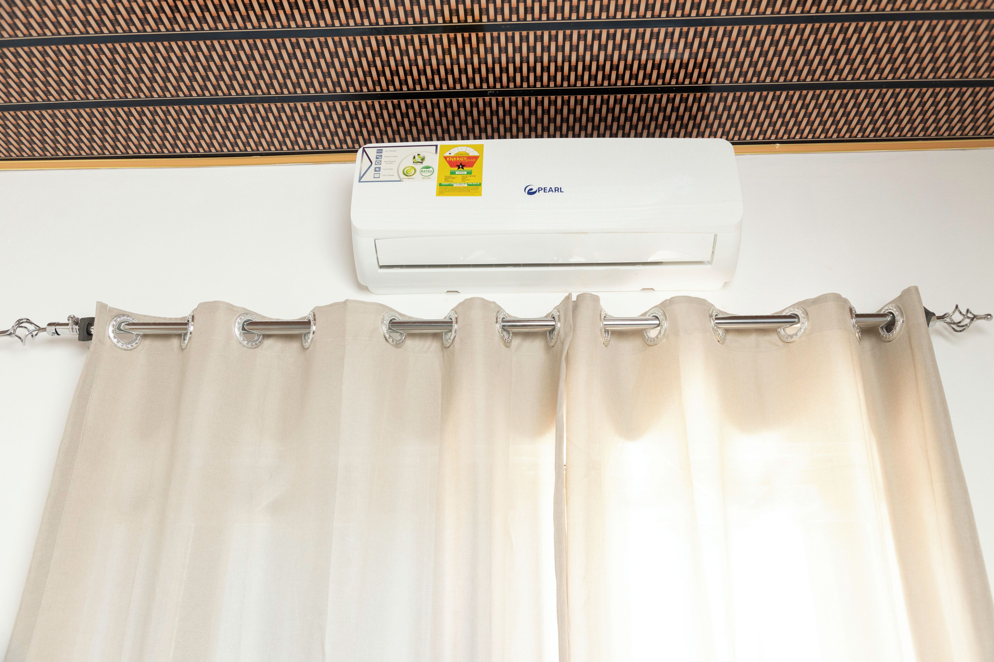 A white air conditioning unit with a label and a control panel, mounted on a wall above a curtain rod with a curtain.