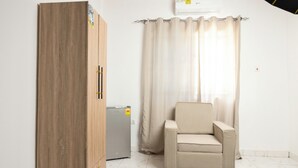 A room with a wooden wardrobe, a small refrigerator, a beige armchair, and a window with curtains.