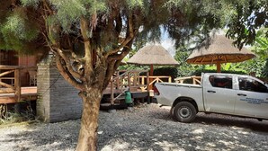 Una camioneta pickup blanca estacionada cerca de una terraza de madera con un paraguas de paja, rodeada de árboles y un edificio de piedra.