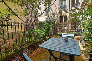 A courtyard with a wooden table and two blue chairs, surrounded by a metal fence and various plants. - La Rochefoulcaud Citycosy (Paris)