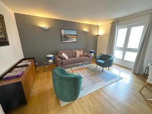A modern living room with a grey sofa, teal armchair, and a coffee table. There is a painting on the wall and a window with curtains. - Appartement Grand Veneur Citycosy (Paris)