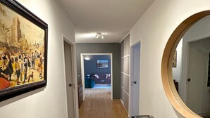 A hallway with wooden flooring, a framed historical painting on the left, a round mirror on the right, and a ceiling light fixture. - Appartement Grand Veneur Citycosy (Paris)