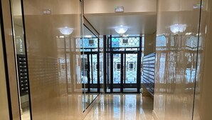 Egy modern, jól megvilágított előtér, csiszolt parkettával, amely egy üvegajtós bejárathoz vezet.