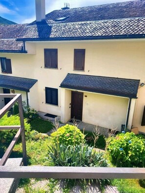 A two-story building with a tiled roof, a small porch, and a garden area with various plants. - LA VILLETTA (Piemonte)