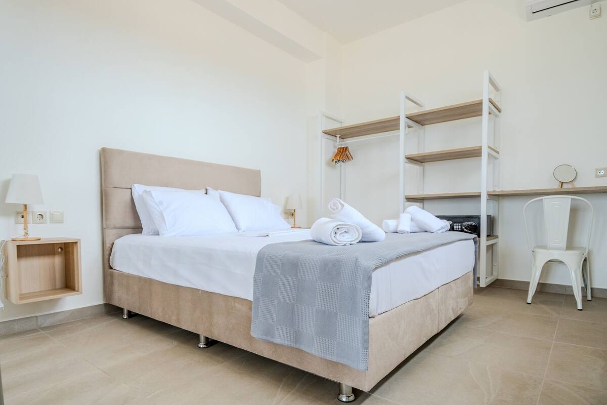 A neatly made bed with a grey blanket, a wooden nightstand with a lamp, and a white chair.