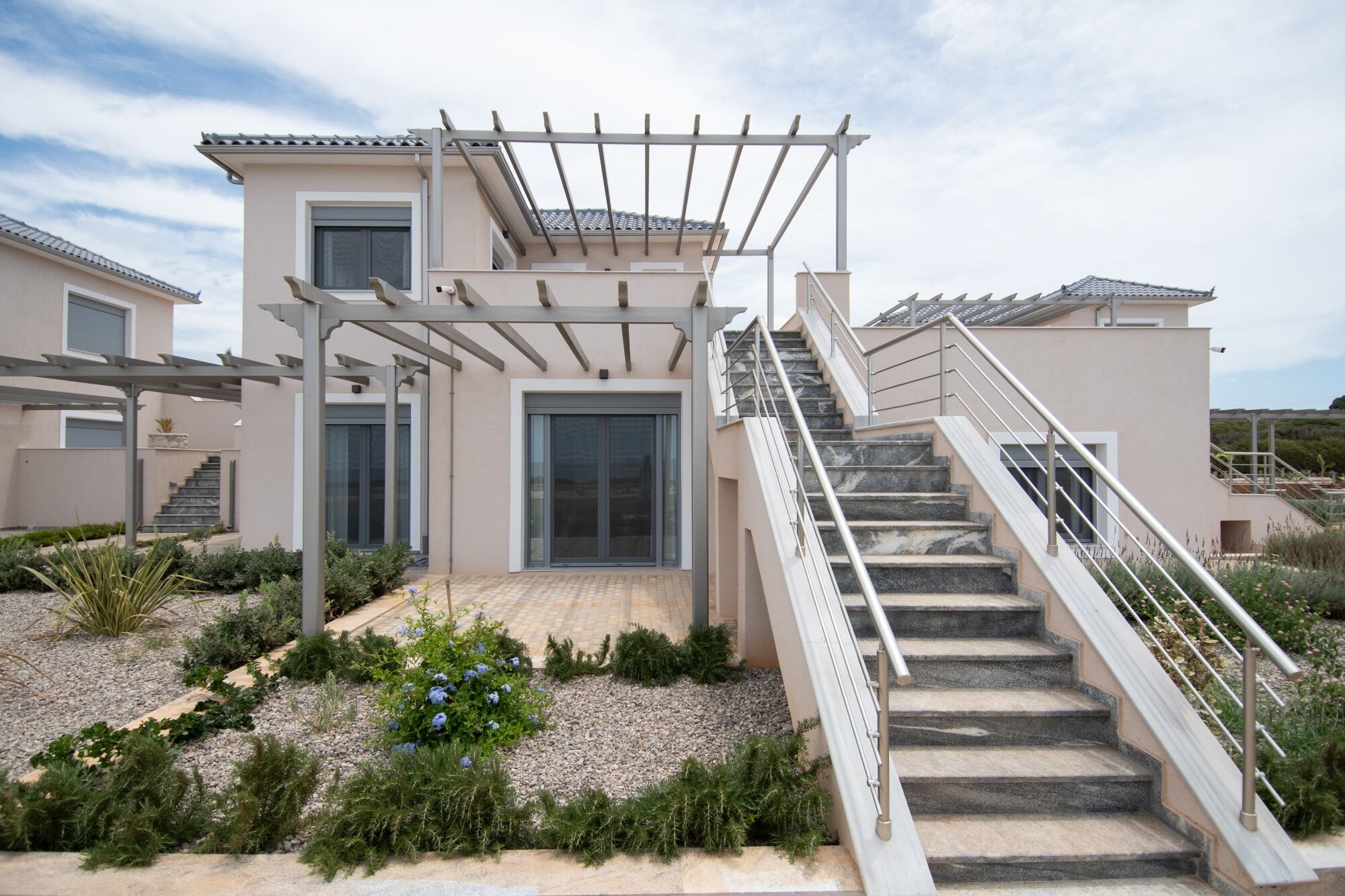 Une maison moderne à deux étages, dotée d’un espace extérieur couvert, d’un escalier et d’un jardin où poussent différentes espèces de plantes.