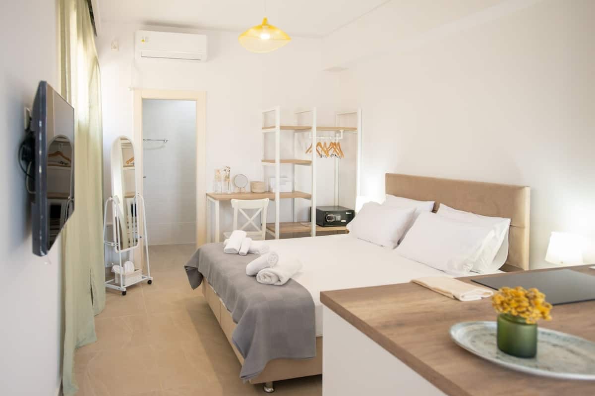 A modern hotel room with a bed, bedside table, and a desk with a chair. There is a TV mounted on the wall, a mirror, and a shelf with toiletries.