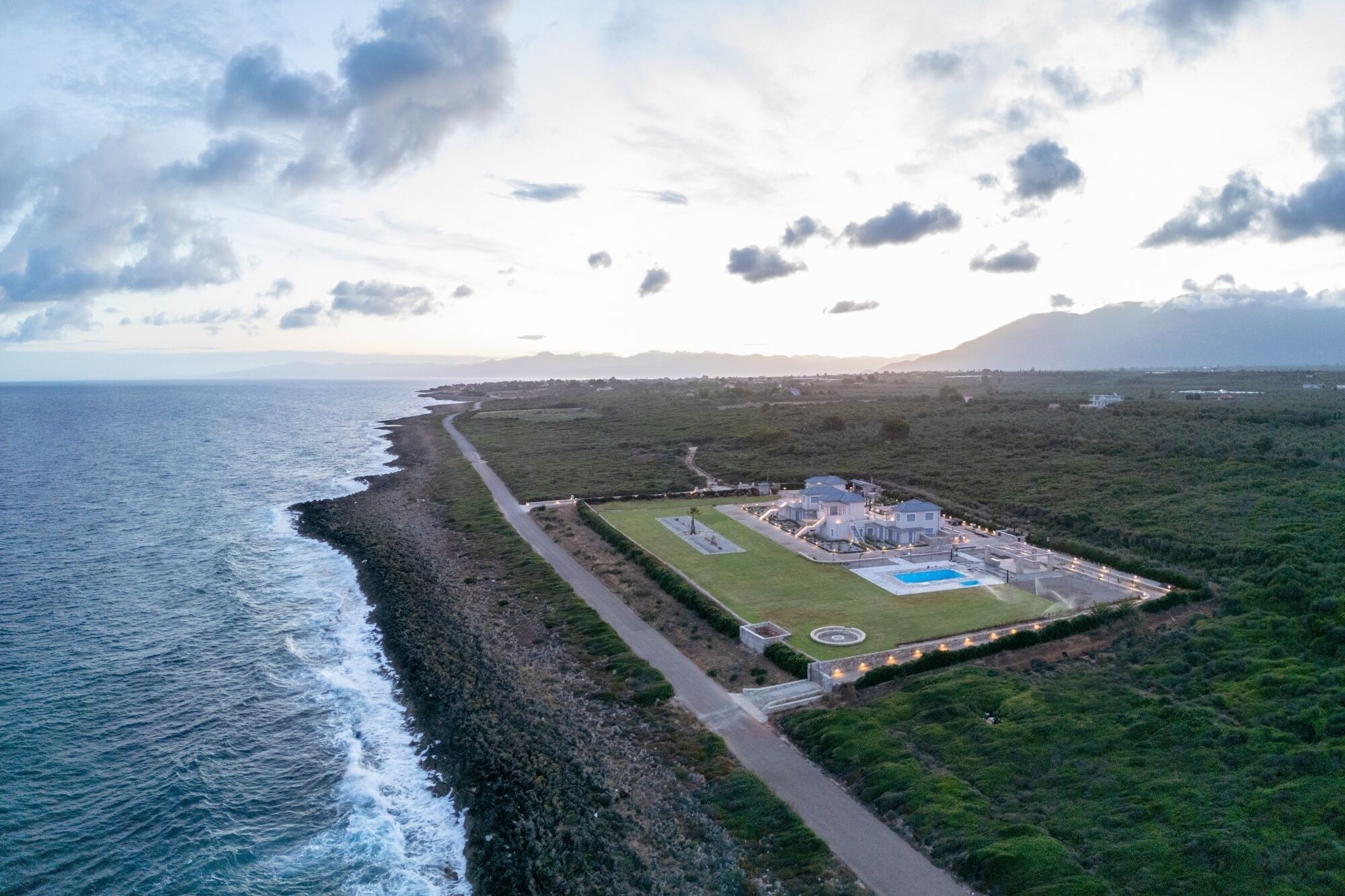 An aerial view of a coastal property with a swimming pool, a paved walkway, and a grassy area.