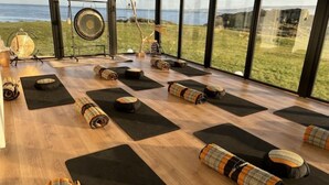 Ein Yoga-Studio mit auf Holzboden angeordneten Matten und Kissen, große Fenster mit Blick auf das Meer.