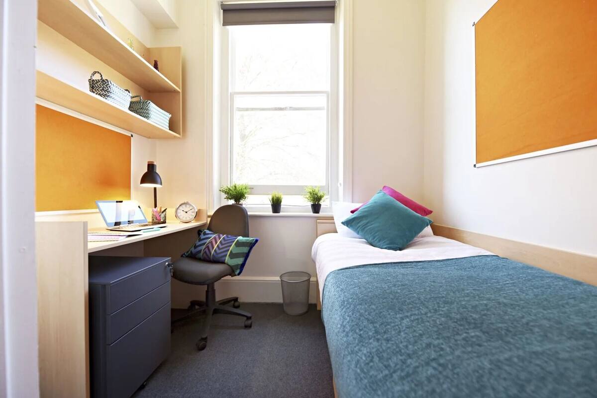 Une chambre à coucher de taille modeste, comprenant un bureau, une chaise et un lit recouvert d’une couverture bleue.