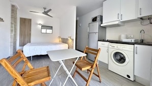A compact room with a bed, a small table, two wooden chairs, a ceiling fan, a washing machine, and a compact kitchenette. - Studio near the beach with terrace AV-76 (ROSES)