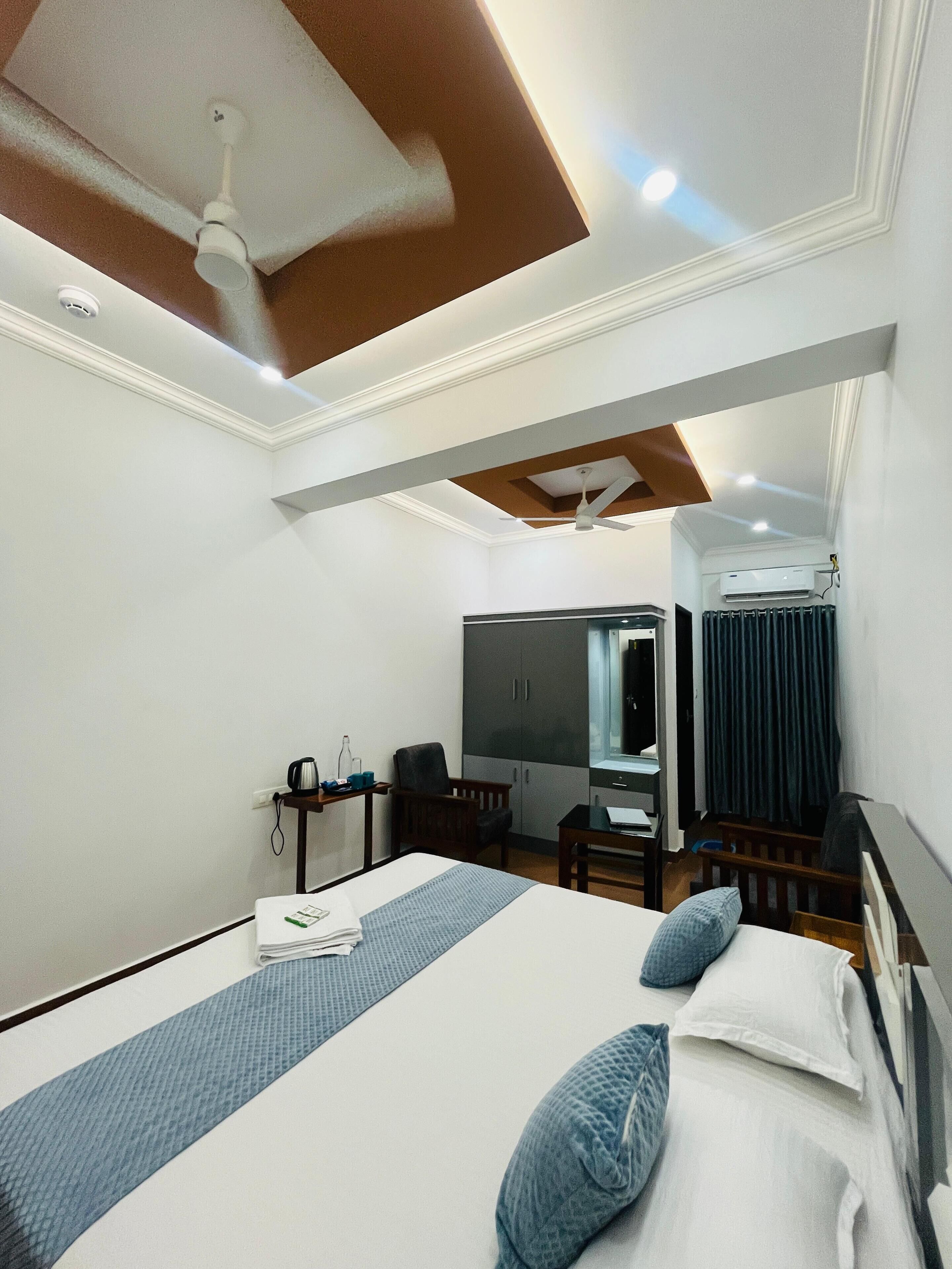 A hotel room with a large bed, a desk with a chair, a wardrobe, a ceiling fan, and an air conditioning unit.