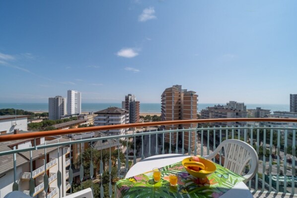 A balcony with a table set for two, overlooking a cityscape and the sea. - Stylish holiday flat in poolside paradise (Lignano Sabbiadoro)