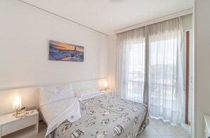 A bedroom with a bed, a nightstand, a lamp, a painting on the wall, and a window with curtains. - Stylish holiday flat in poolside paradise (Lignano Sabbiadoro)