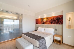 A modern bedroom with a large bed, a nightstand, a wall-mounted lamp, and a view of a glass door leading to a balcony.